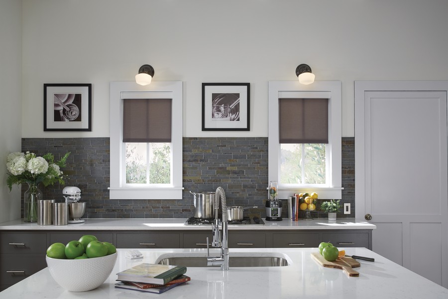 Daylight flows in through two windows over a kitchen counter, illuminating the counter and center cabinet featuring a sink, bowl of fruit and notebooks.