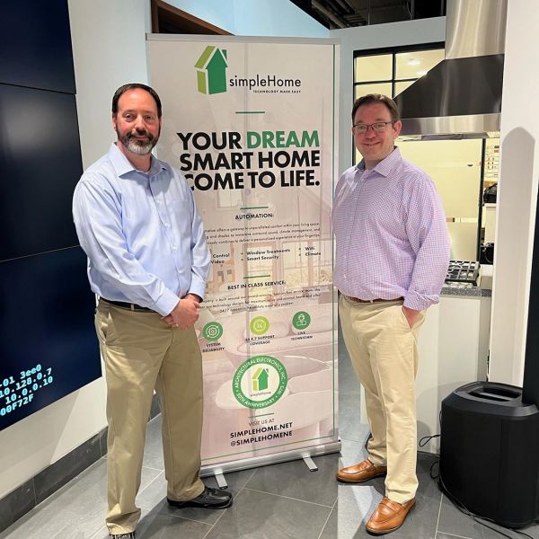 Two men standing indoors next to a promotional banner, both smiling at the camera.