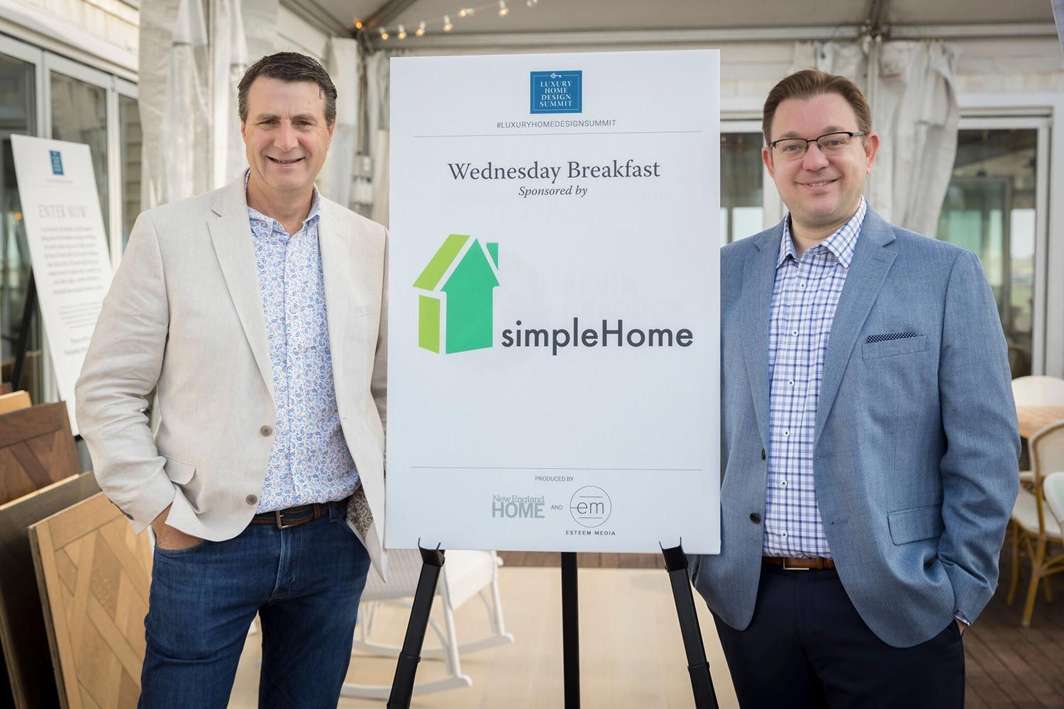 Two men standing next to a sign for a Wednesday breakfast event in a bright, outdoor setting.