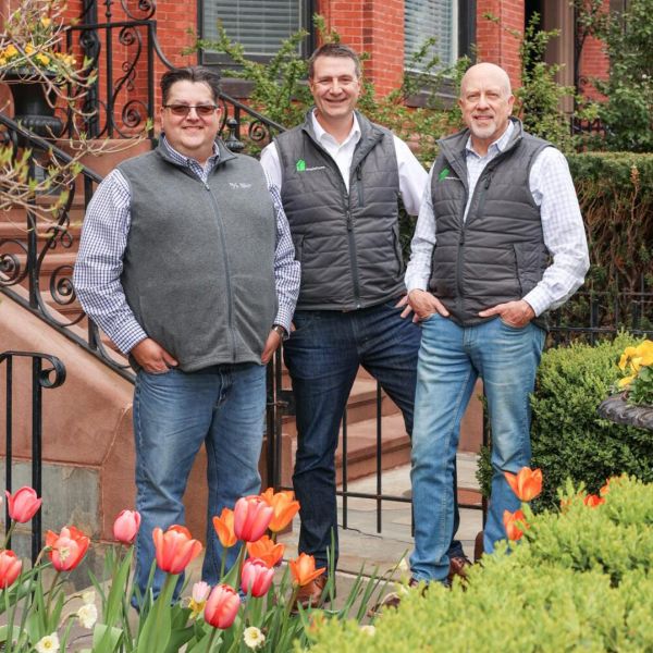 Three men in matching vests posing in front of a red brick building and a garden with tulips.